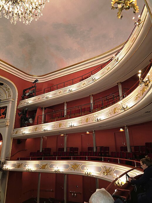 Innenraum eines Theaters mit mehreren geschwungenen Rängen. Die Wände sind rot, die Verzierungen weiß und gold. Auf den Balkonen stehen Reihen von roten Sitzplätzen. Mehrere Kronleuchter hängen von der Decke, die mit einer gemalten, wolkigen Szene gestaltet ist.