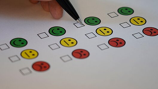 Das Bild zeigt einen Zettel mit positiven, neutralen und negativen Smileys auf einem Umfragebogen sowie eine Hand mit Stift.
